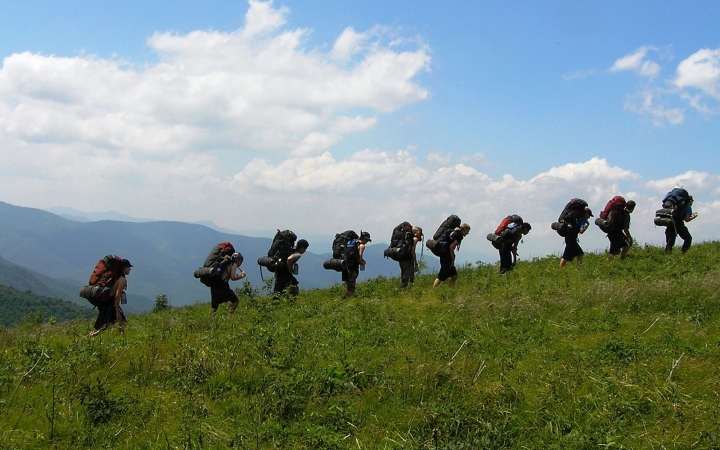 A line of people wearing backpacks hike in a line across a grassy meadow under blue skies. There are mountains in the background. 